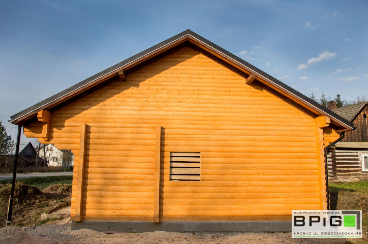 Żółty dom z bali w surowym stanie, z widocznym zadaszeniem, małym oknem z deskami i fundamentami, na tle błękitnego nieba i innych budynków.