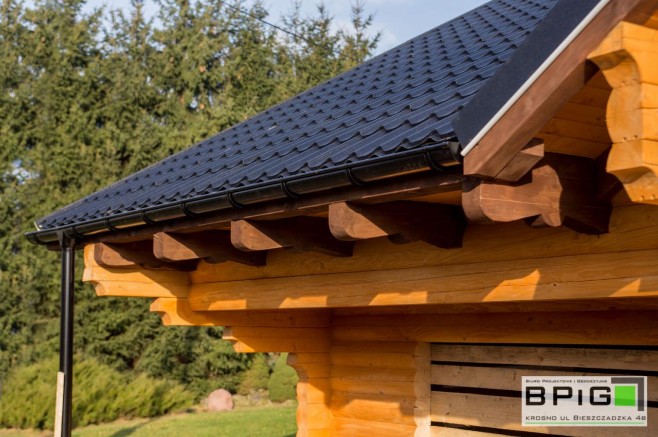 Fragment domu z bali z ciemną, metalową dachówką i czarnymi rynnami, widoczny ozdobny okap i logo firmy na ścianie.