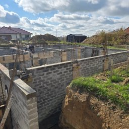 Firma Remontowo Budowlana HOP- home our passion - Budowa fundamentów z bloczków betonowych, widoczne zbrojenie pionowe i szalunki, dwóch pracowników w trakcie pracy, w tle domy jednorodzinne i niebo z chmurami.