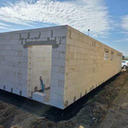 Firma Remontowo Budowlana HOP- home our passion - Wznoszenie ścian domu z białych bloczków, widoczny otwór drzwiowy i okienne, izolacja fundamentów w czarnym kolorze, teren wokół wyrównany, błękitne niebo w tle.