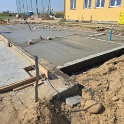 Firma Remontowo Budowlana HOP- home our passion - Wylewanie betonowej płyty fundamentowej z widocznymi elementami szalunku, pionową rurą instalacyjną i pracującym robotnikiem w tle, na placu budowy z żółtym budynkiem i stertą materiałów izolacyjnych.