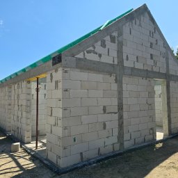 Firma Remontowo Budowlana HOP- home our passion - Surowy stan domu murowanego z betonu komórkowego Ytong, z widocznymi betonowymi nadprożami i wieńcem, zabezpieczony zieloną siatką, na tle błękitnego nieba.