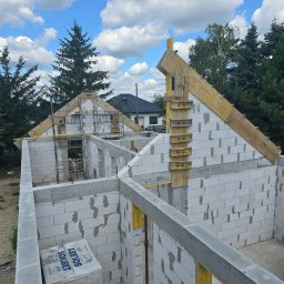 Firma Remontowo Budowlana HOP- home our passion - Wznoszenie ścian domu z białych bloczków Solbet, widoczne drewniane szalunki dachowe, w tle zielone drzewa i błękitne niebo z chmurami, murarz pracuje na zewnątrz.