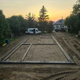 Przygotowany zarys fundamentów domu jednorodzinnego z widocznymi wystającymi prętami zbrojeniowymi, na tle zachodzącego słońca i zabudowy jednorodzinnej.