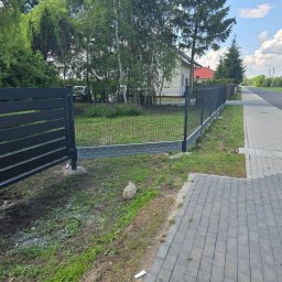 Firma Remontowo Budowlana HOP- home our passion - Nowoczesne, grafitowe ogrodzenie panelowe z bramą wjazdową, częściowo zamontowane, z widocznymi betonowymi podstawami słupków, na tle zielonego trawnika i domu jednorodzinnego.