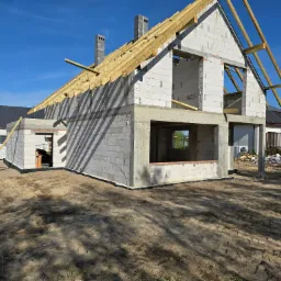 Dwukondygnacyjny budynek w stanie surowym otwartym z białych bloczków, z drewnianą konstrukcją dachu, widoczne dwa kominy, teren wokół budynku piaszczysty.