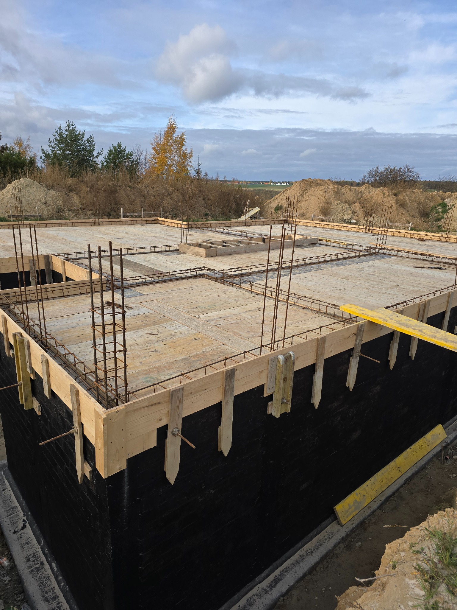 Zabezpieczone czarną izolacją fundamenty z drewnianym szalunkiem i zbrojeniem, widok z góry na plac budowy z piaszczystym nasypem w tle.