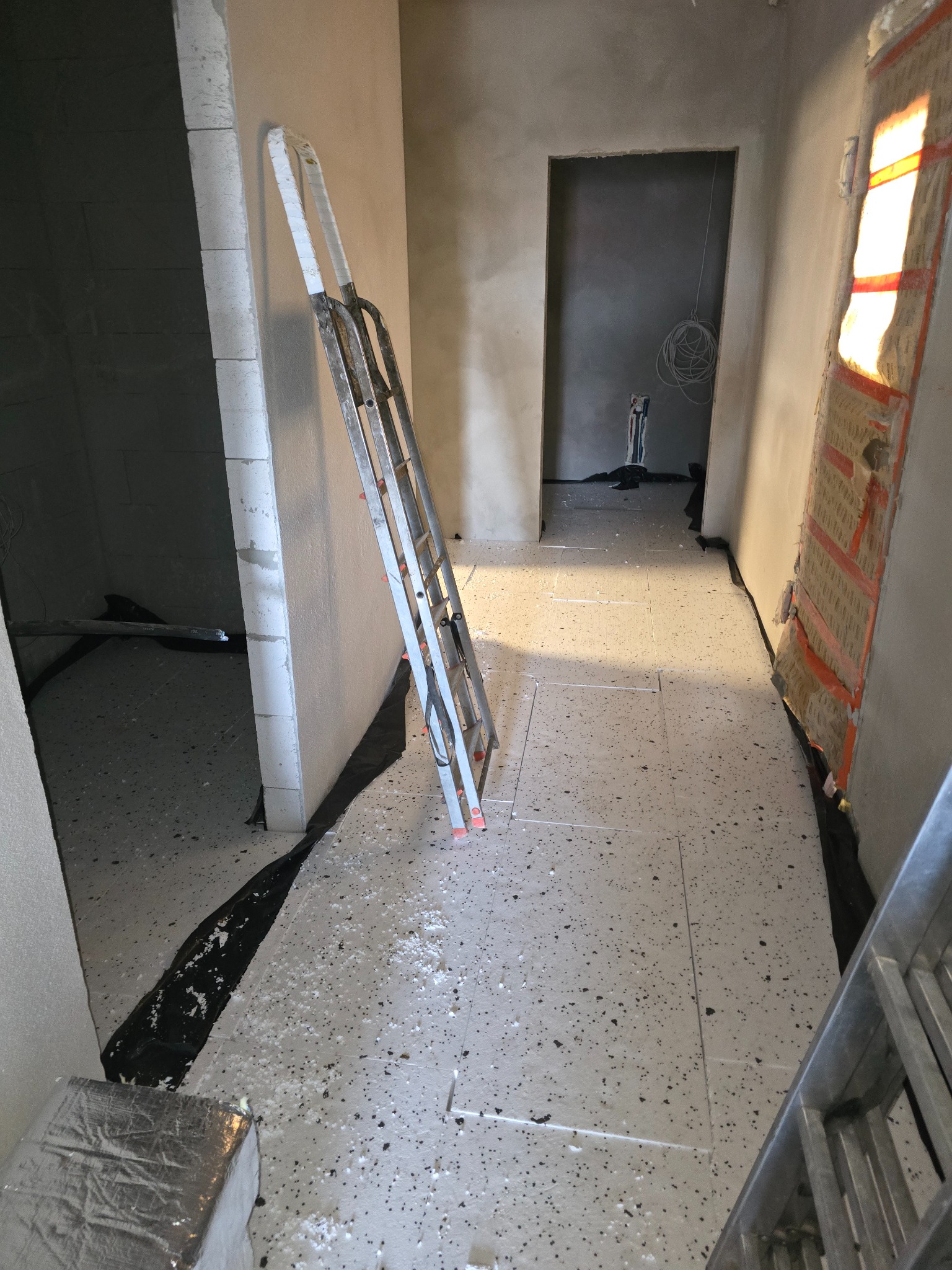 Wnętrze w trakcie prac wykończeniowych: ściany surowe, widoczna drabina aluminiowa oparta o ścianę, podłoga wyłożona płytkami lastryko, fragment izolacji ściany widoczny w tle, zabezpieczenie folią...