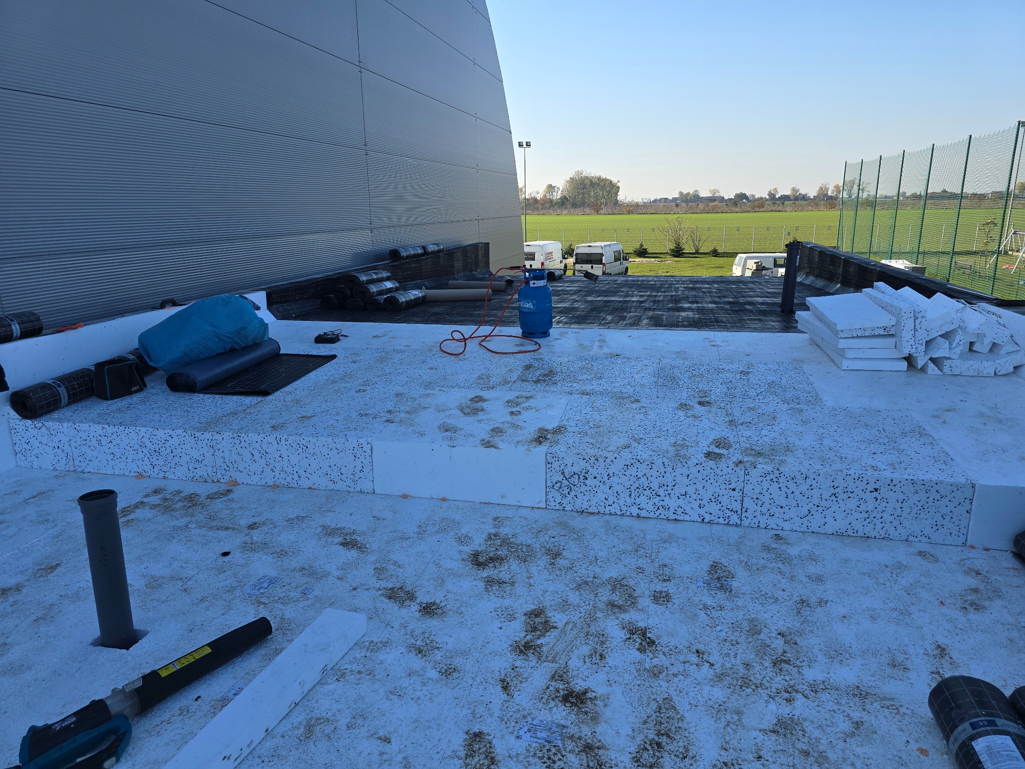 Izolacja dachu płaskiego styropianem z widocznymi rolkami papy, butlą gazową i dmuchawą do czyszczenia, na tle budynku przemysłowego i zielonego krajobrazu.