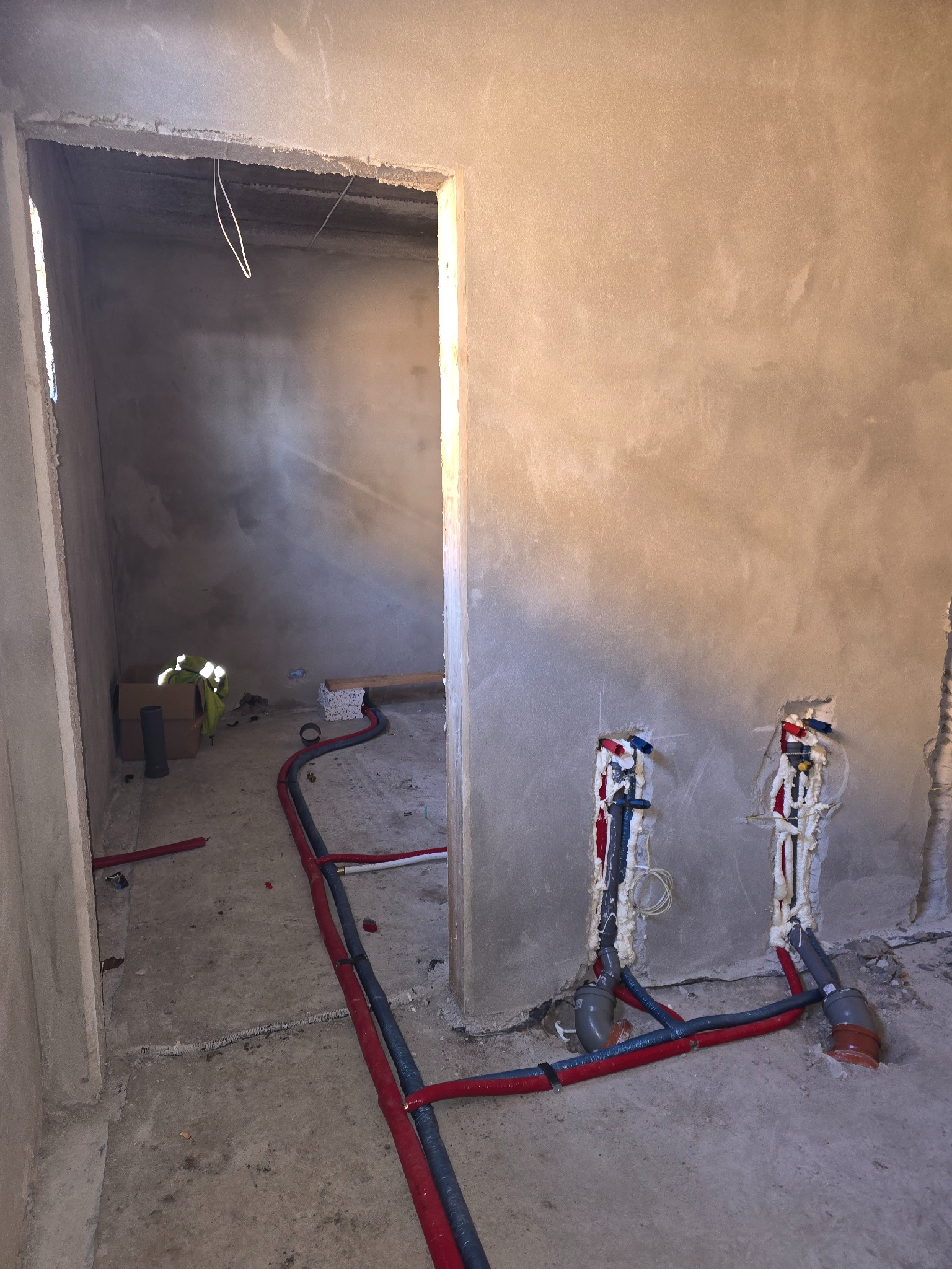 Wnętrze w trakcie remontu: surowe ściany pokryte tynkiem, wystające rury instalacji wodnej zabezpieczone pianką montażową, widoczne przewody w podłodze, w tle otwarte przejście do kolejnego...