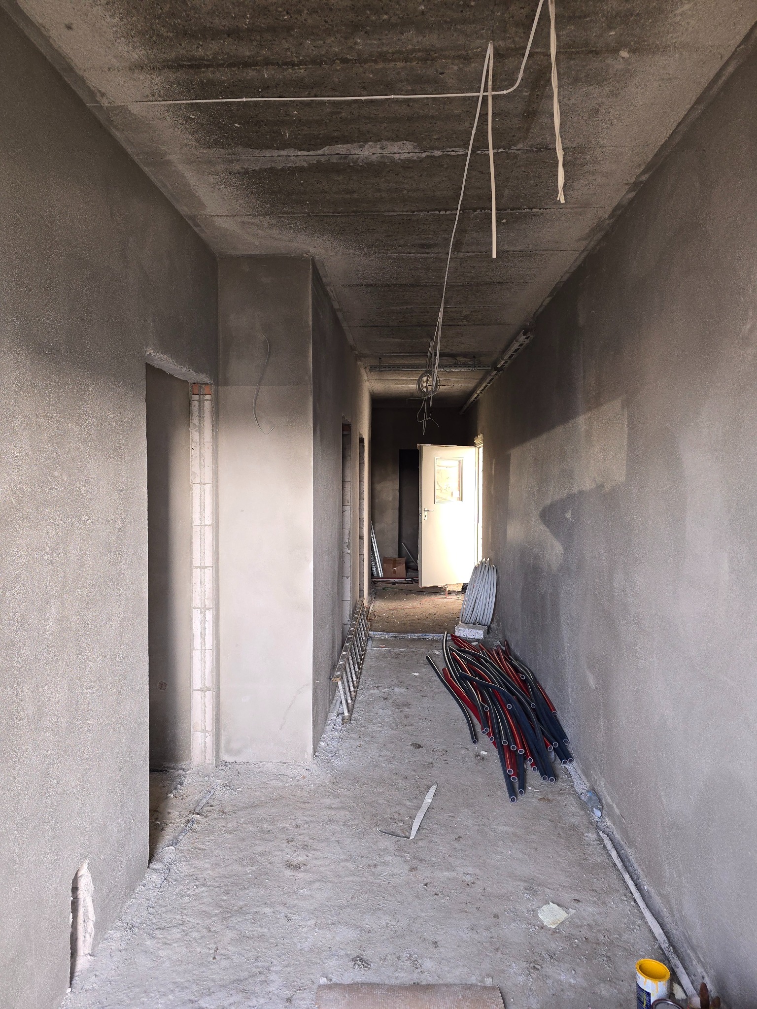 Wnętrze korytarza w stanie surowym, ściany pokryte szarym tynkiem, widoczne przewody elektryczne, drabina oparta o ścianę i materiały budowlane na podłodze.