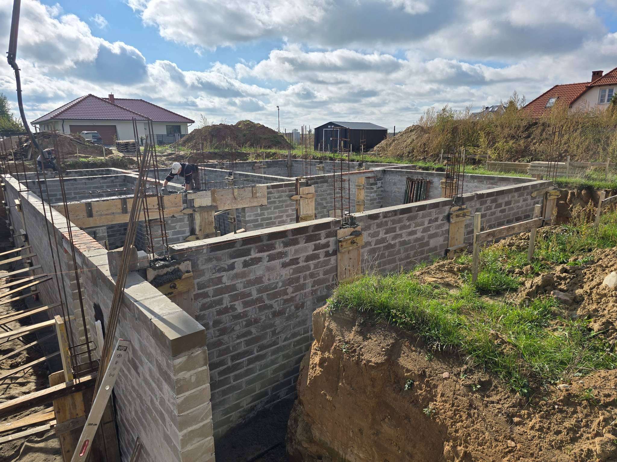 Budowa fundamentów z bloczków betonowych, widoczne zbrojenie pionowe i szalunki, dwóch pracowników w trakcie pracy, w tle domy jednorodzinne i niebo z chmurami.