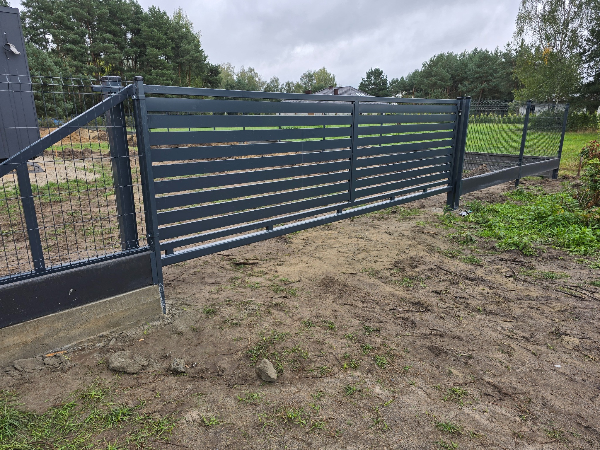 Nowoczesna, przesuwna brama w kolorze antracytowym z poziomymi panelami, zamontowana na betonowej podmurówce, z widoczną siatką ogrodzeniową po bokach i drzewami w tle.