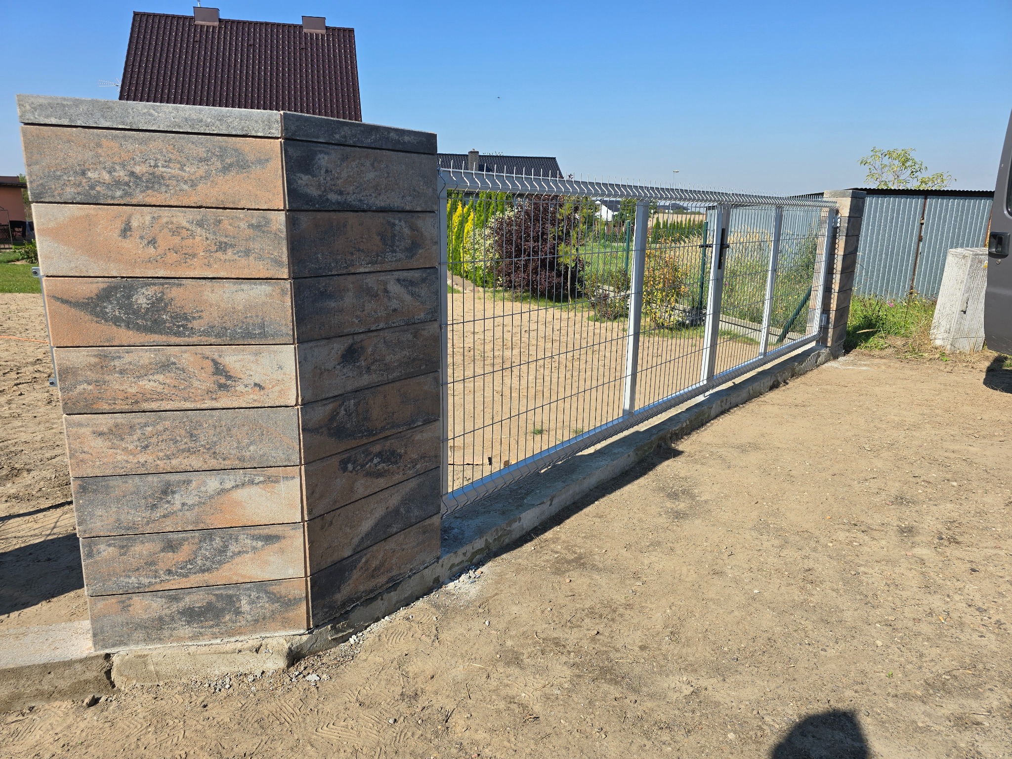 Nowoczesne ogrodzenie panelowe z metalową siatką i słupkami z bloczków betonowych o nieregularnym wzorze, widok z zewnątrz posesji, w tle dom z brązowym dachem i roślinność.