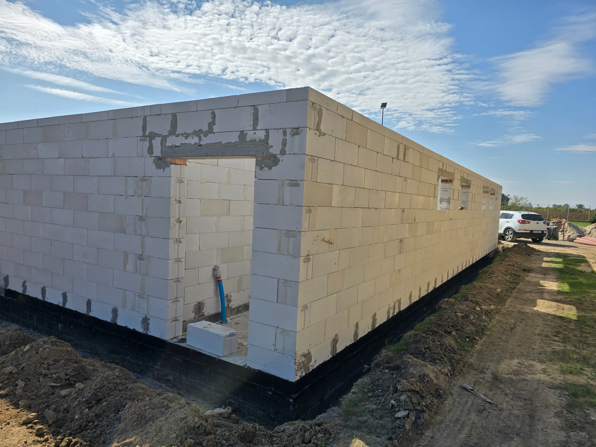 Wznoszenie ścian domu z białych bloczków, widoczny otwór drzwiowy i okienne, izolacja fundamentów w czarnym kolorze, teren wokół wyrównany, błękitne niebo w tle.