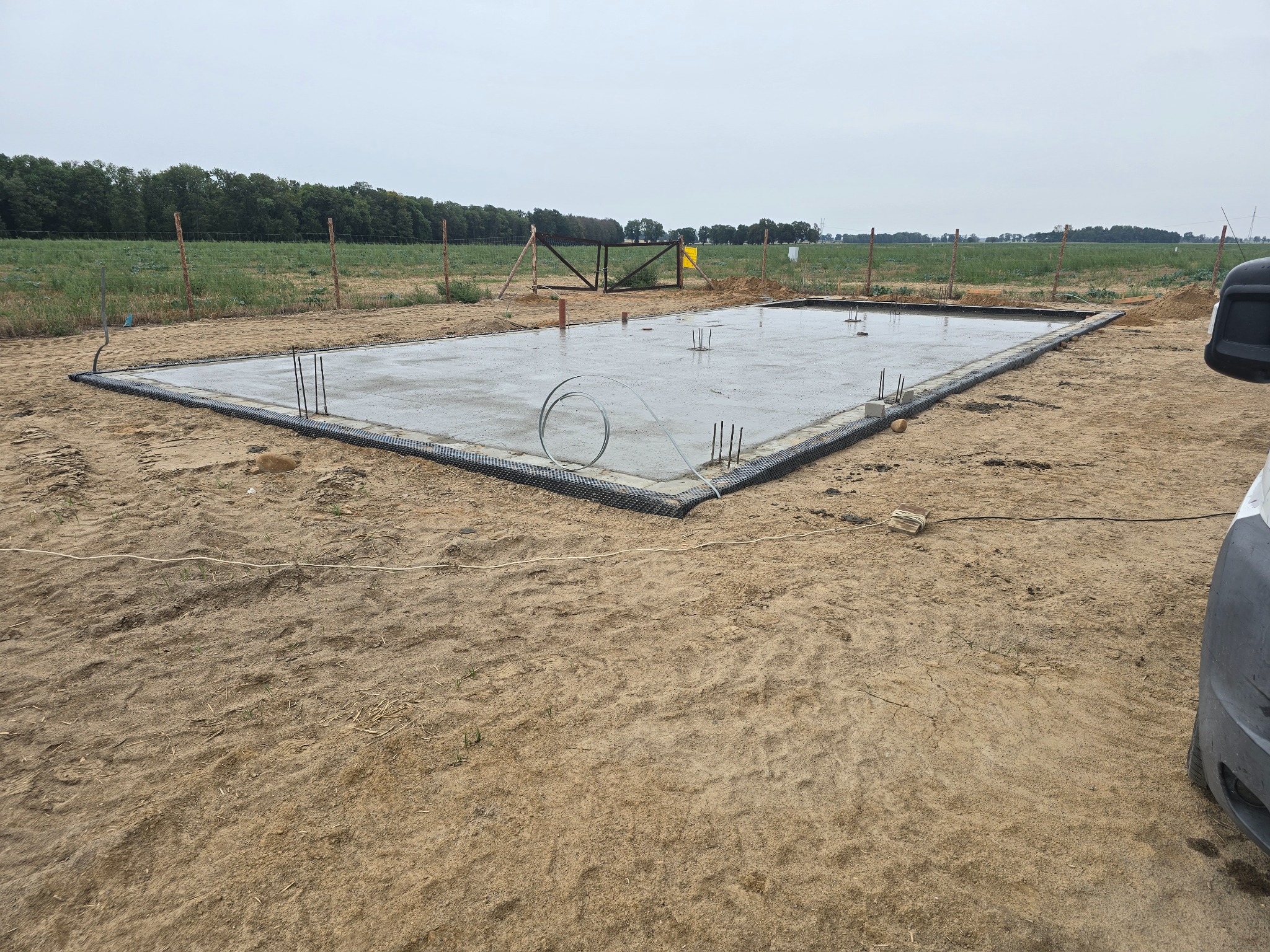 Wylany fundament z wystającymi prętami zbrojeniowymi, zabezpieczony czarną folią, otoczony piaszczystym terenem i ogrodzeniem z drewnianych słupków. W tle widoczne pole i drzewa.
