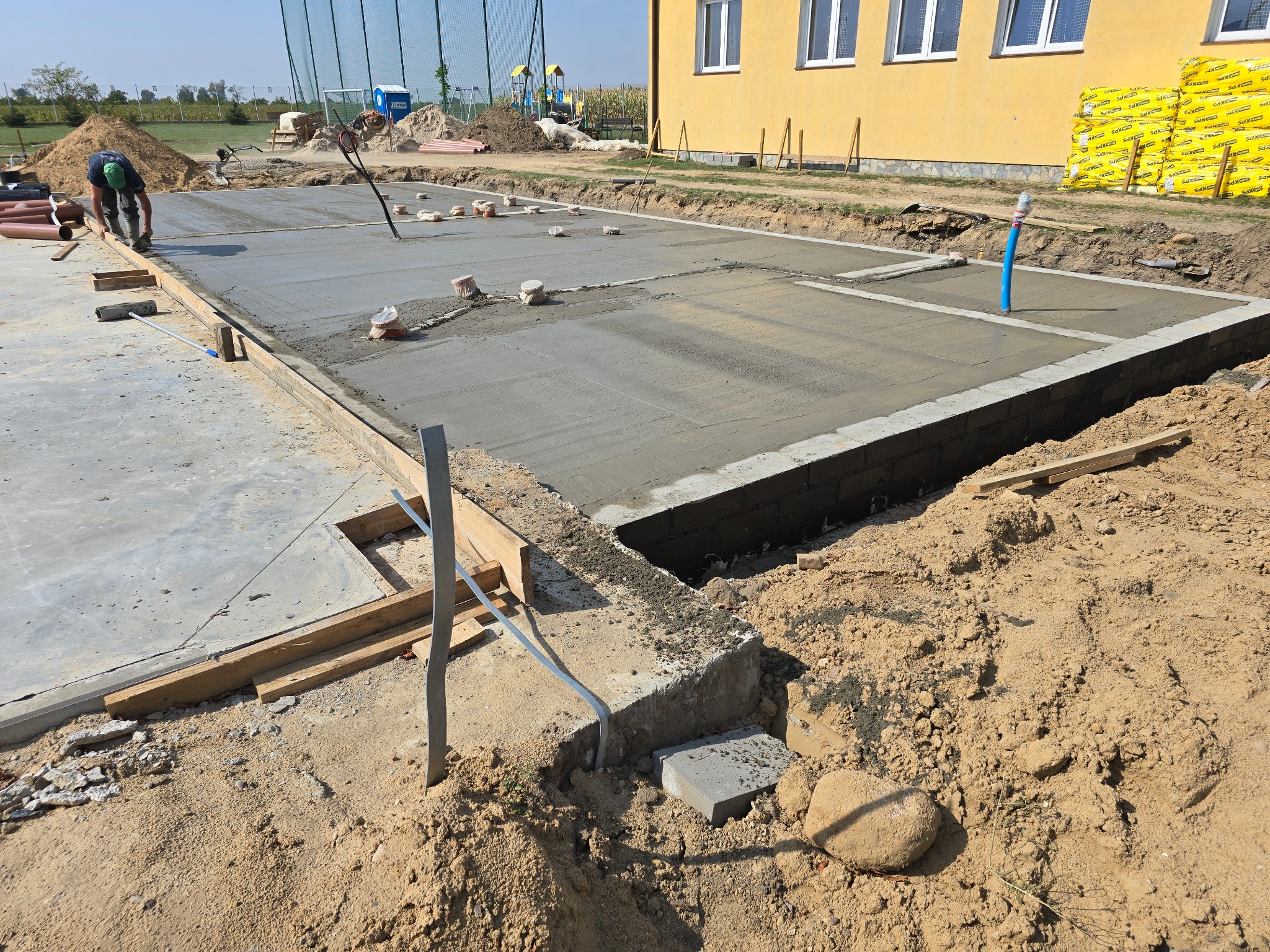 Wylewanie betonowej płyty fundamentowej z widocznymi elementami szalunku, pionową rurą instalacyjną i pracującym robotnikiem w tle, na placu budowy z żółtym budynkiem i stertą materiałów izolacyjnych.