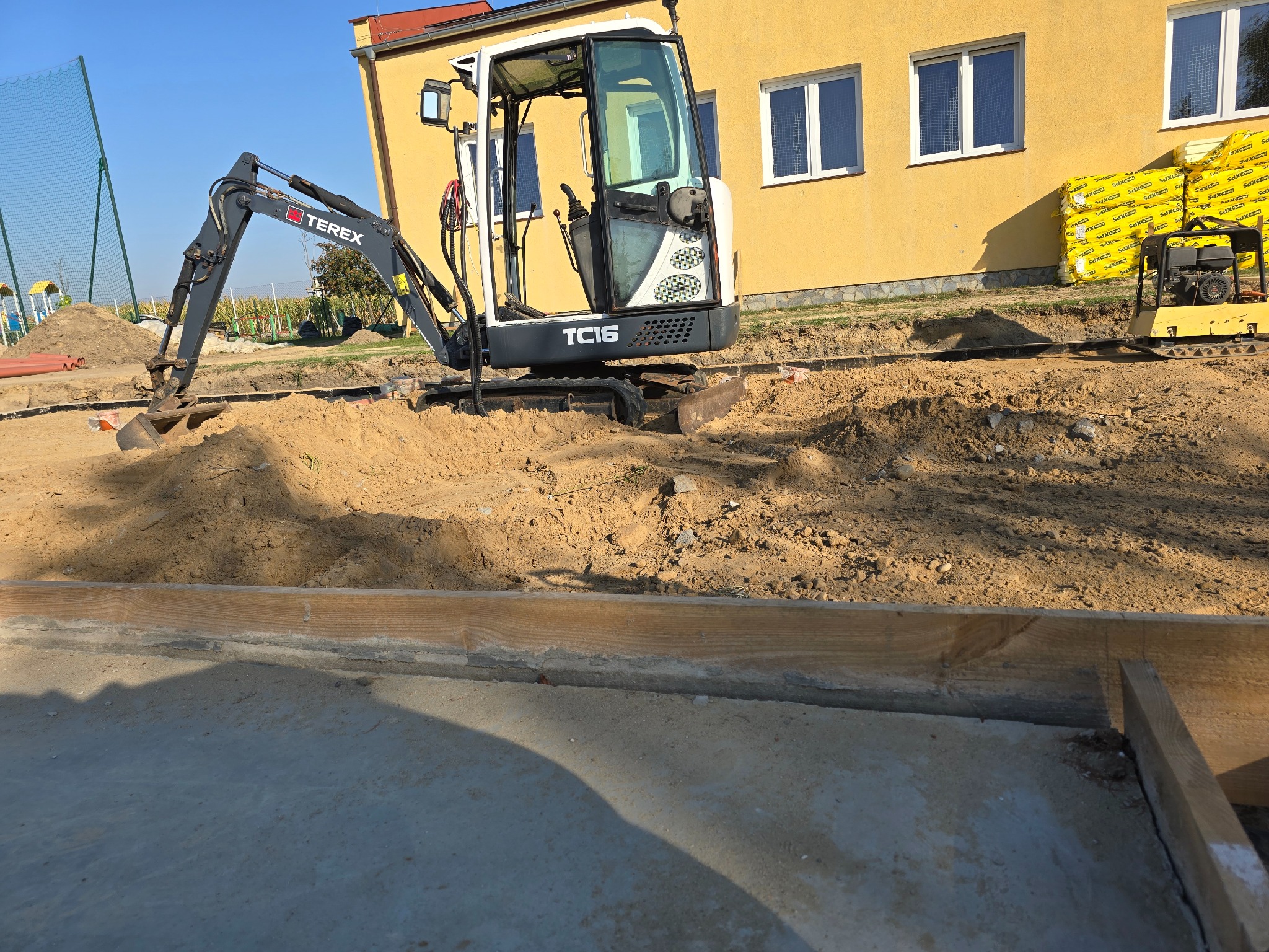Mini koparka Terex TC16 podczas niwelacji terenu pod budowę fundamentów w Kłecku. W tle widoczny budynek z oknami i materiały izolacyjne.