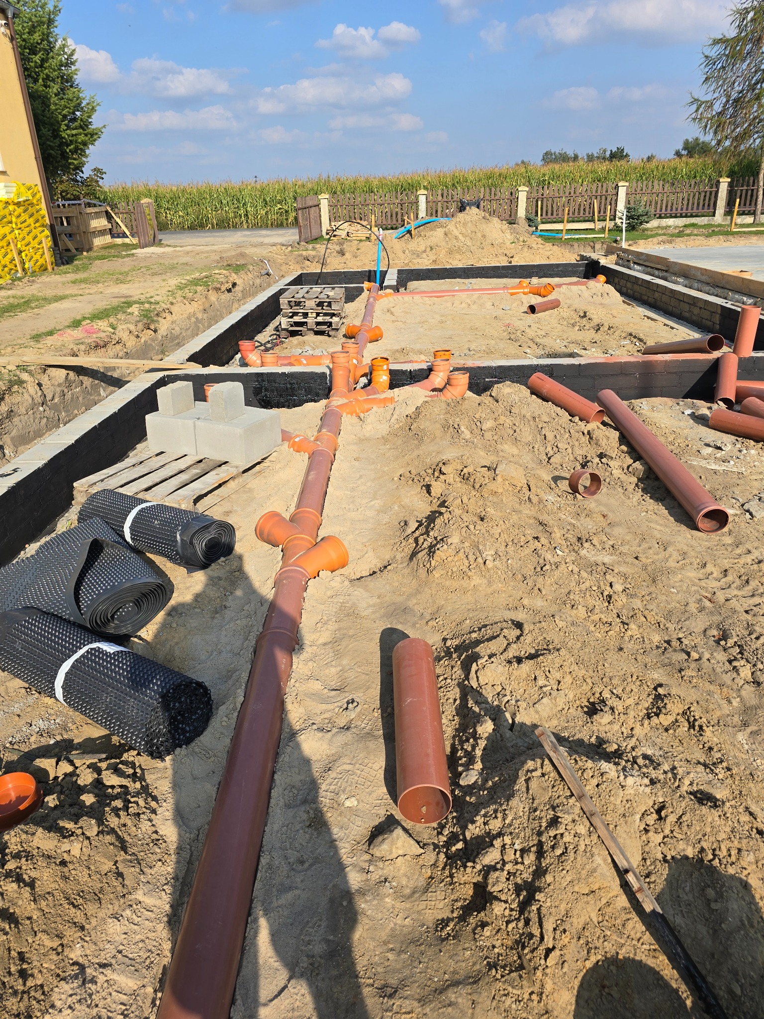 Fundamenty domu w trakcie budowy z widocznymi ścianami fundamentowymi zabezpieczonymi izolacją, ułożonymi rurami kanalizacyjnymi, rolkami folii kubełkowej i stertą piasku w słoneczny dzień.