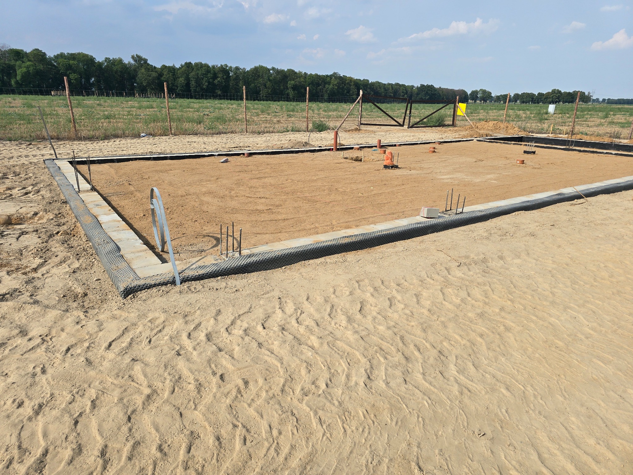 Przygotowany teren pod fundamenty z widoczną izolacją pionową i wystającymi prętami zbrojeniowymi, otoczony piaszczystym terenem i polami uprawnymi.