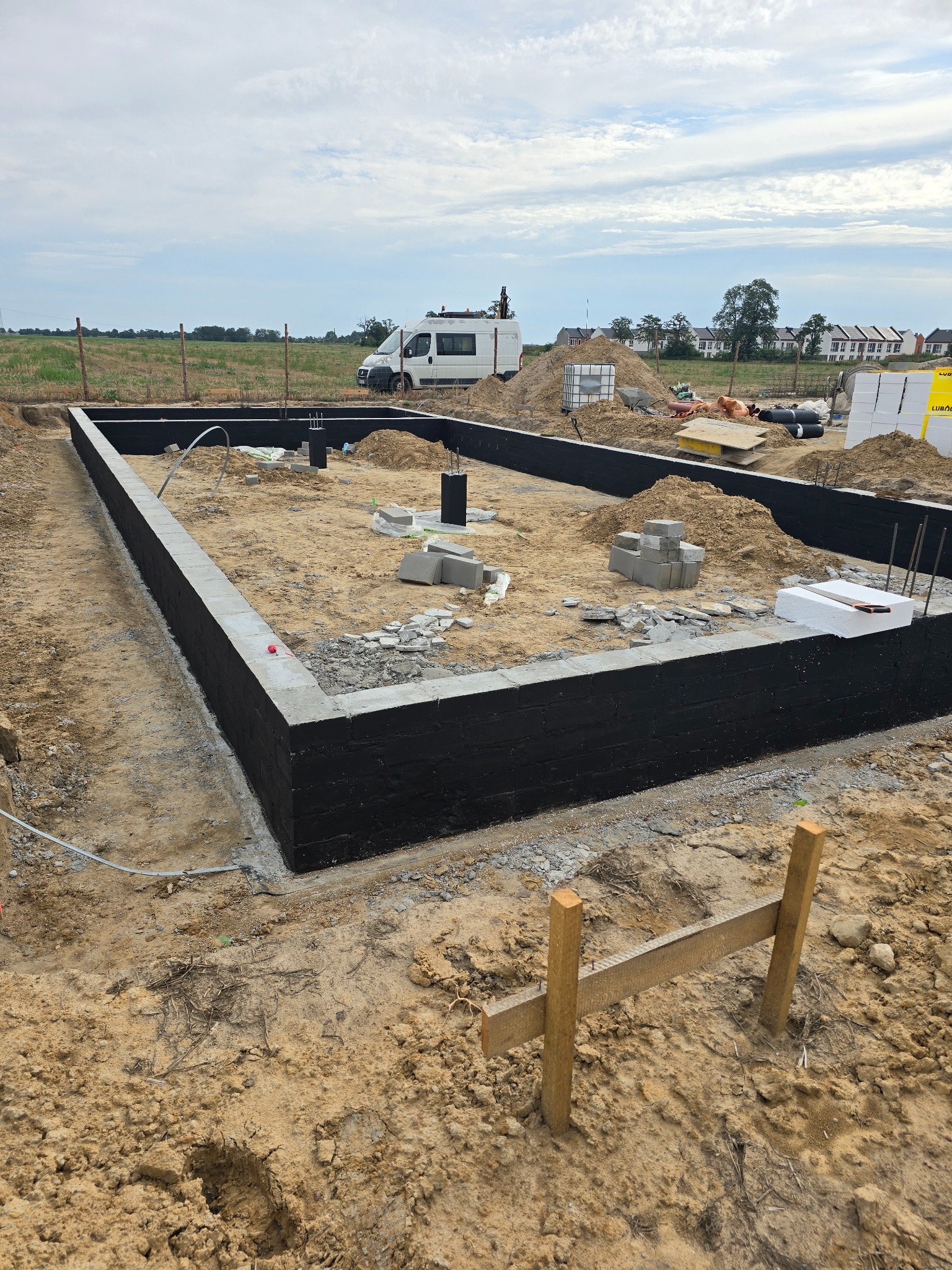 Fundamenty domu jednorodzinnego z izolacją przeciwwilgociową, widoczny teren budowy, materiały budowlane i samochód dostawczy w tle.
