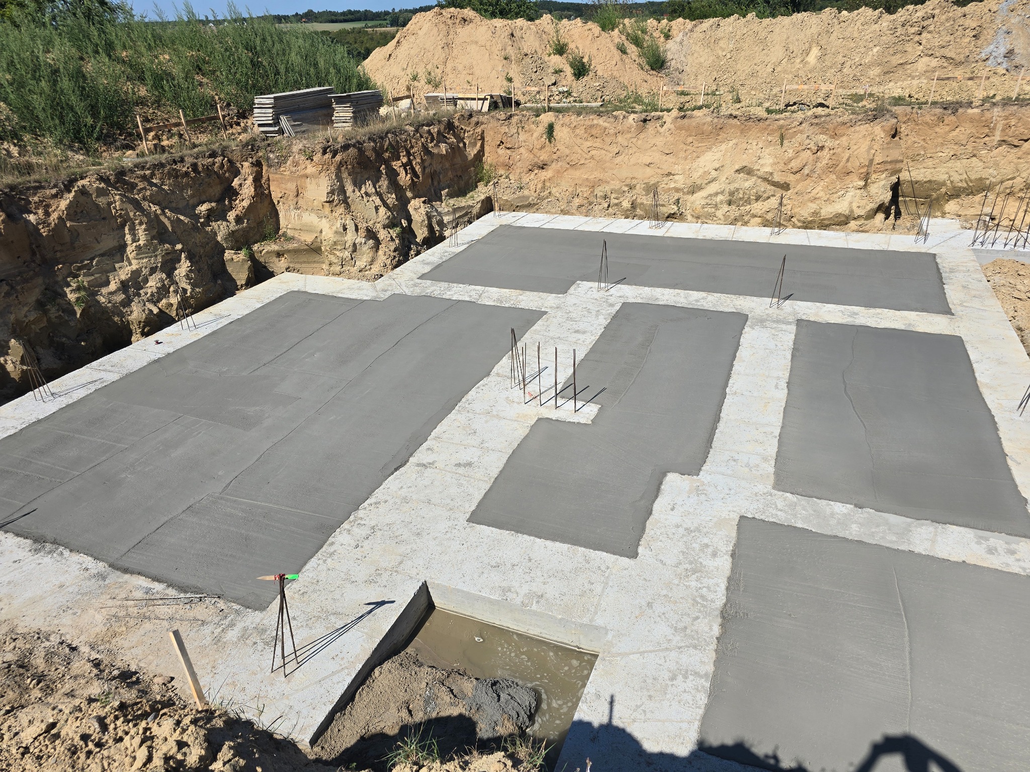 Wylane fundamenty z wystającymi prętami zbrojeniowymi, widok z góry na wykop, z boku sterta desek szalunkowych.