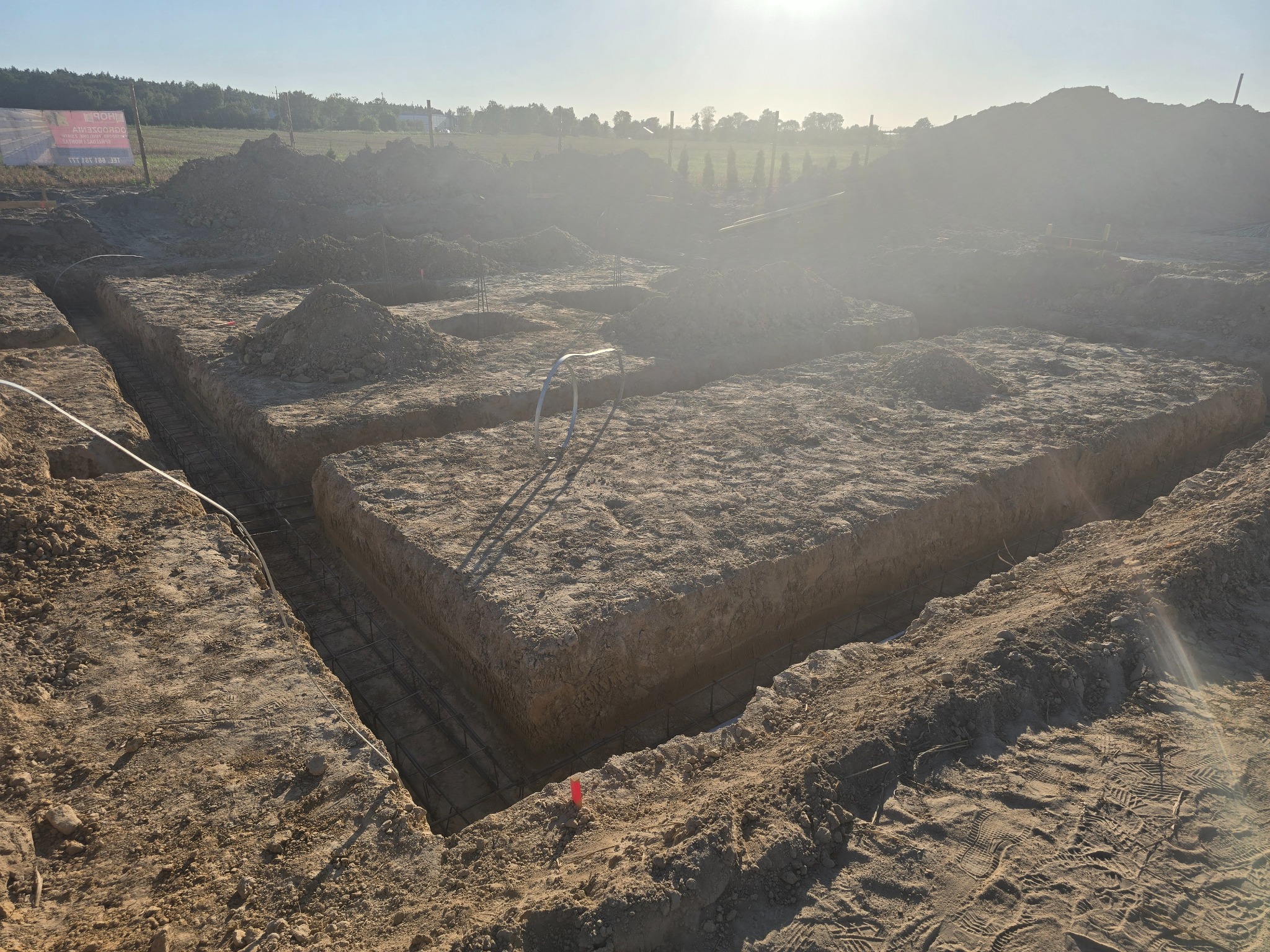 Wykonane wykopy pod fundamenty z widocznym zbrojeniem, hałdami ziemi i odległym lasem w tle, oświetlone silnym słońcem.