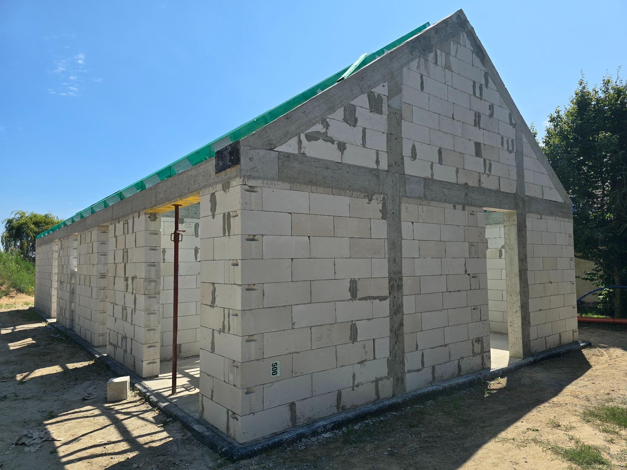Surowy stan domu murowanego z betonu komórkowego Ytong, z widocznymi betonowymi nadprożami i wieńcem, zabezpieczony zieloną siatką, na tle błękitnego nieba.