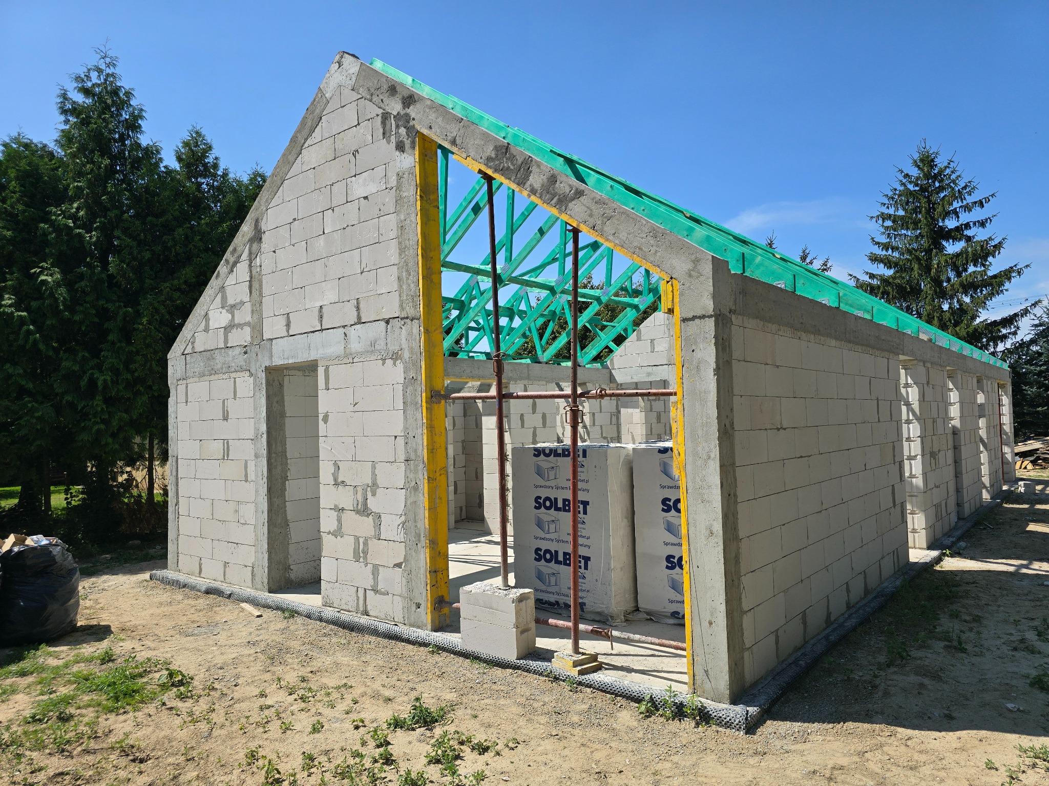 Niedokończony budynek z betonu komórkowego z widocznym szalunkiem i zieloną konstrukcją dachu, na zewnątrz leżą bloczki Solbet