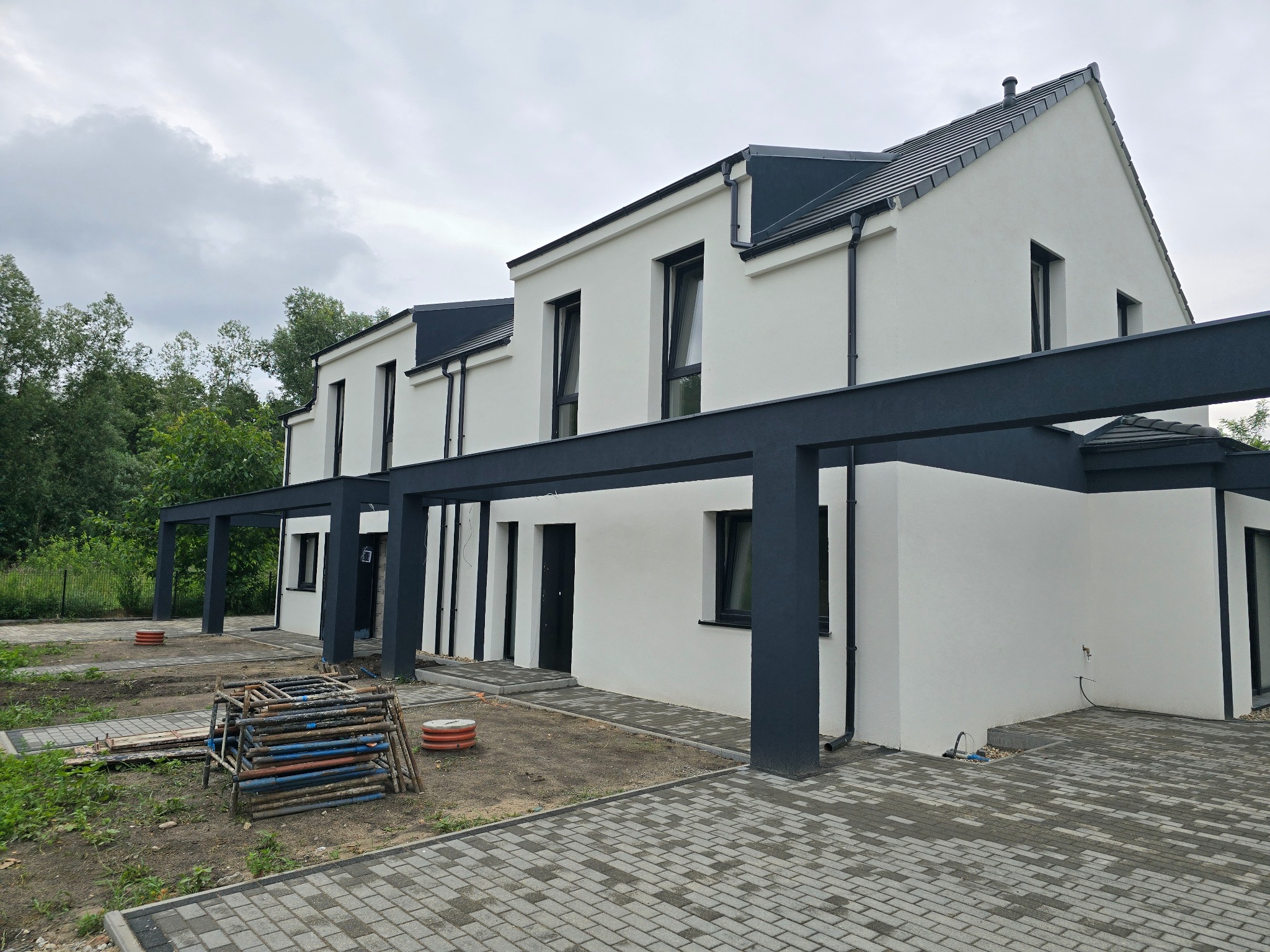 Nowoczesny, dwukondygnacyjny dom z białymi ścianami i grafitowymi elementami architektonicznymi, takimi jak okna, dach i konstrukcja wspornikowa nad wejściem. Na pierwszym planie ułożona kostka...