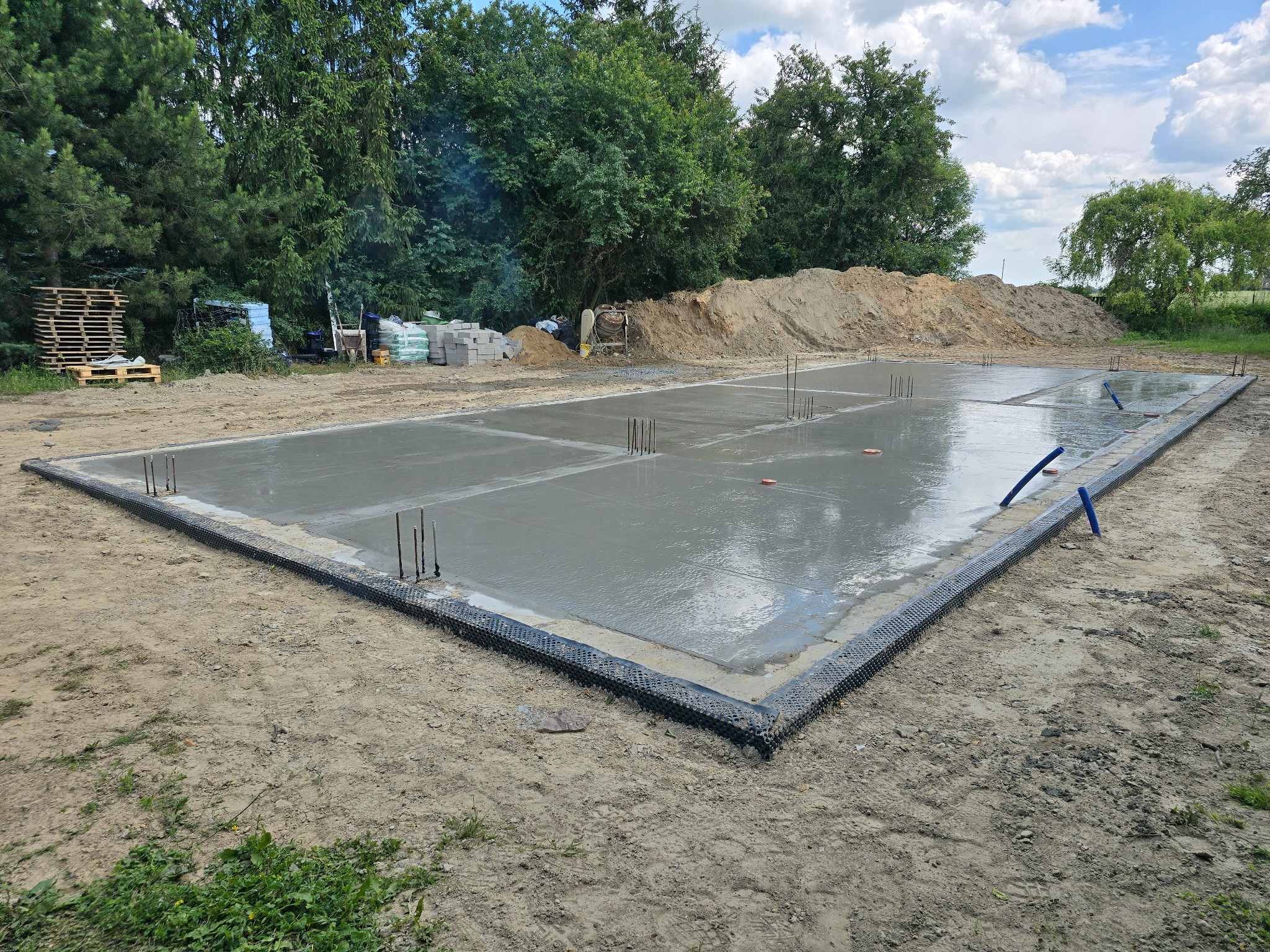 Świeżo wylana, mokra płyta fundamentowa z wystającymi prętami zbrojeniowymi, otoczona czarną folią kubełkową, na piaszczystym gruncie, z hałdą ziemi i zielenią w tle.