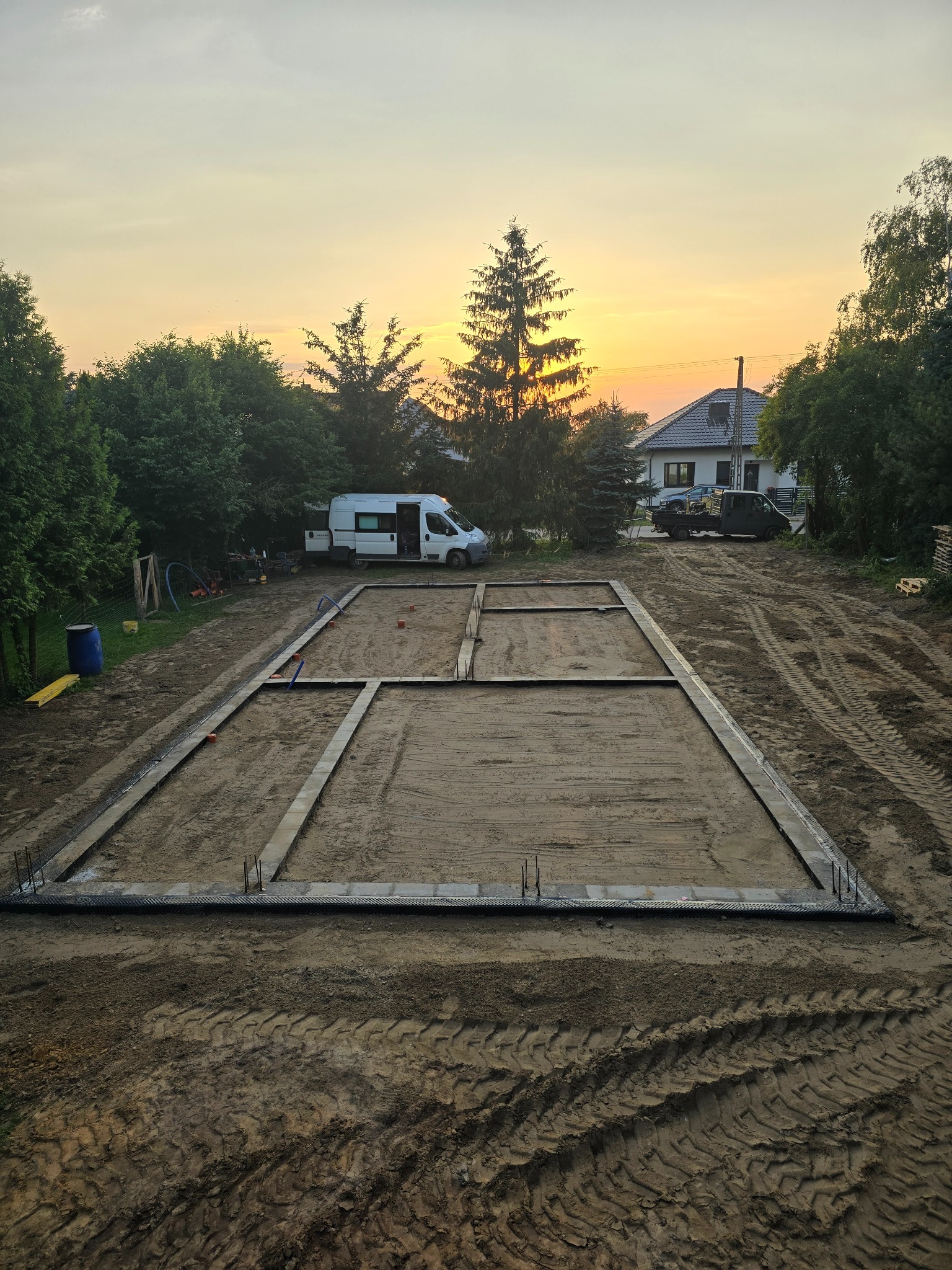 Przygotowany zarys fundamentów domu jednorodzinnego z widocznymi wystającymi prętami zbrojeniowymi, na tle zachodzącego słońca i zabudowy jednorodzinnej.