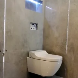 Nowoczesna, podwieszana toaleta w łazience z szarą, betonową okładziną ścian i podłogi. W ścianie wnęka z podświetleniem LED i otwór montażowy na przycisk spłukujący.