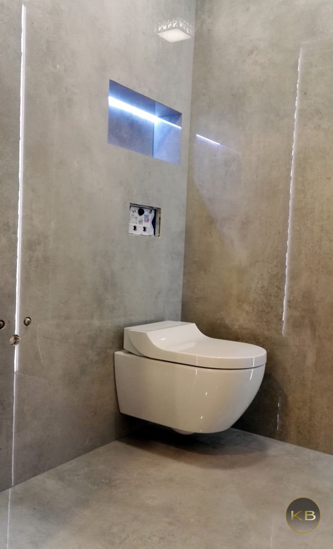 Nowoczesna, podwieszana toaleta w łazience z szarą, betonową okładziną ścian i podłogi. W ścianie wnęka z podświetleniem LED i otwór montażowy na przycisk spłukujący.