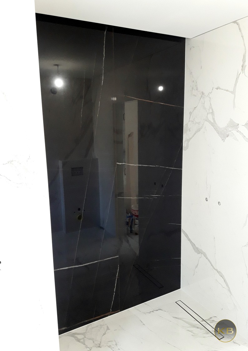 Częściowo wykończona łazienka z czarnymi płytami imitującymi marmur na ścianie, kontrastującymi z białymi płytami na sąsiedniej ścianie i podłodze, widoczny odpływ liniowy w podłodze.