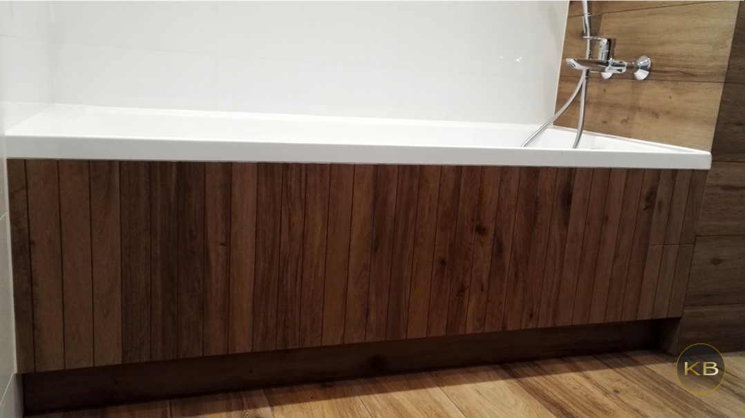 Biała wanna z obudową z pionowych paneli imitujących ciemne drewno, chromowana bateria, płytki imitujące drewno na ścianie i podłodze.