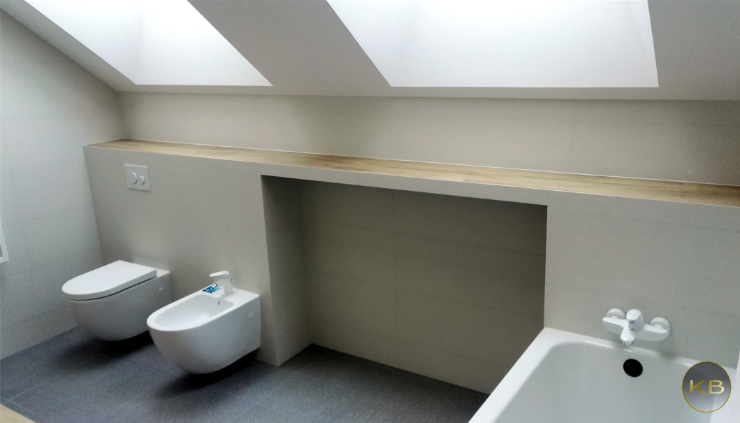 Jasna łazienka na poddaszu ze skośnym sufitem i oknami dachowymi, wisząca toaleta i bidet, wanna z baterią oraz półka z drewnianym blatem.