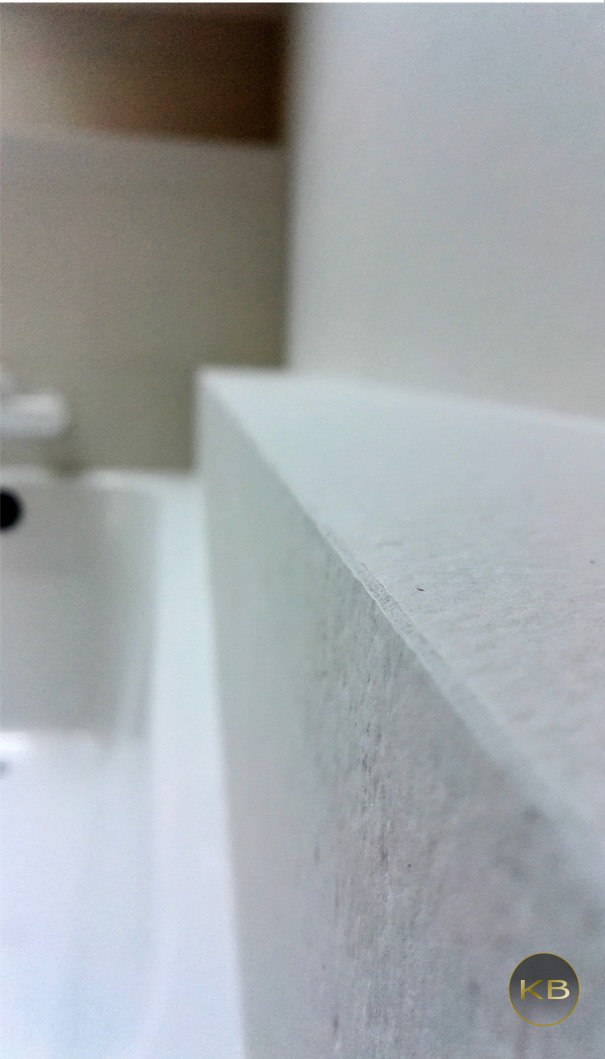 Bliski kadr na krawędź białego blatu umywalki w łazience, widoczna umywalka w tle, logo firmy w prawym dolnym rogu.