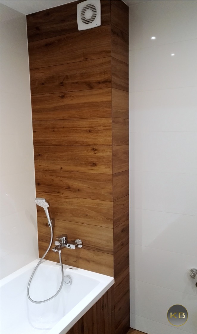 Nowoczesna łazienka z wanną obudowaną płytkami imitującymi drewno, wentylator na ścianie, białe płytki na ścianach, chromowana bateria wannowa z słuchawką.