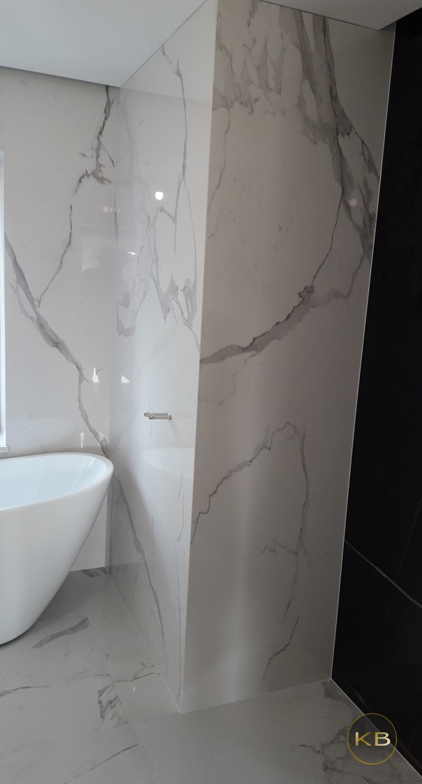 Nowoczesna łazienka z białą, wolnostojącą wanną i ścianami wyłożonymi wielkoformatowymi płytkami imitującymi marmur Carrara, z widocznym fragmentem czarnej ściany kabiny prysznicowej.