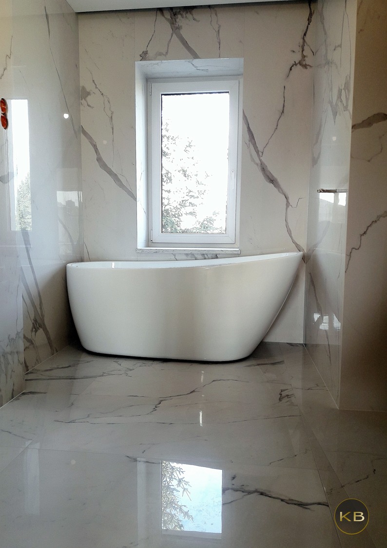 Nowoczesna łazienka z wolnostojącą wanną i marmurowymi płytkami, okno nad wanną odbija widok drzew.