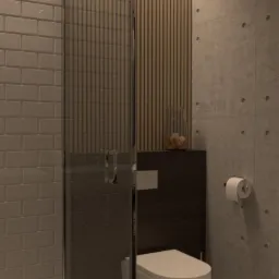 Nowoczesna łazienka z kabiną prysznicową, podwieszaną toaletą i betonową ścianą z dekoracyjnymi otworami, uzupełniona o drewniane panele i szklany wazon z dekoracją.