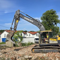 Wyburzenia Pomorze sp.zoo - Żółto-szara koparka Volvo na gruzowisku, w tle zabudowa mieszkalna i błękitne niebo. Operator w kabinie. Widoczne fragmenty zburzonej konstrukcji i cegieł.