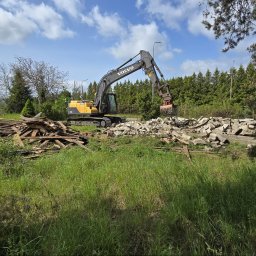 Wyburzenia Pomorze sp.zoo - Żółto-szara koparka Volvo z chwytakiem hydraulicznym usuwa gruz i drewno z terenu zielonego, na tle drzew i błękitnego nieba.