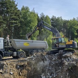 Wyburzenia Pomorze sp.zoo - Żółto-czarna koparka Volvo ładuje gruz do szarej ciężarówki MAN Wan-Dróg na tle zieleni drzew. Operator nadzoruje proces.