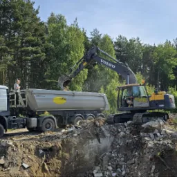 Żółto-szara koparka Volvo EC160DL załadowuje gruz do ciężarówki MAN z pracownikiem na pace, na tle lasu i nieba.