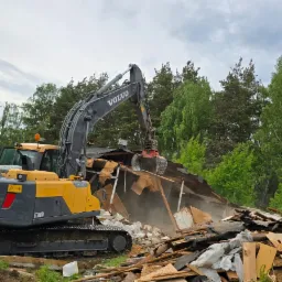 Żółto-szara koparka Volvo EC160DL w trakcie rozbiórki drewnianego budynku. Wokół gruz i drzewa, pochmurne niebo. Widoczny etap prac rozbiórkowych.