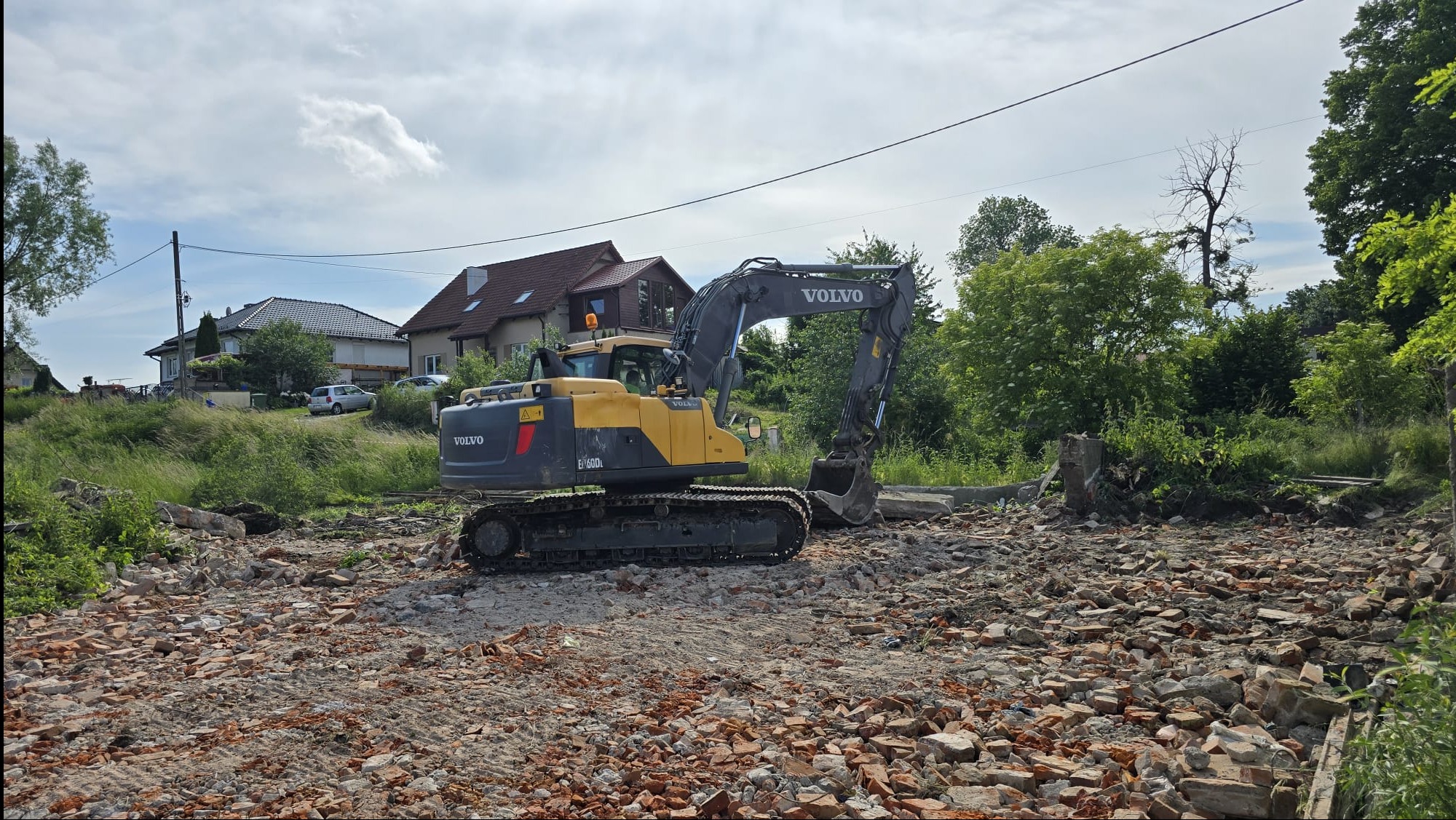 Żółta koparka Volvo na terenie po rozbiórce, z gruzem ceglanym. W tle zabudowa jednorodzinna i drzewa. Słoneczny dzień, błękitne niebo z chmurami.