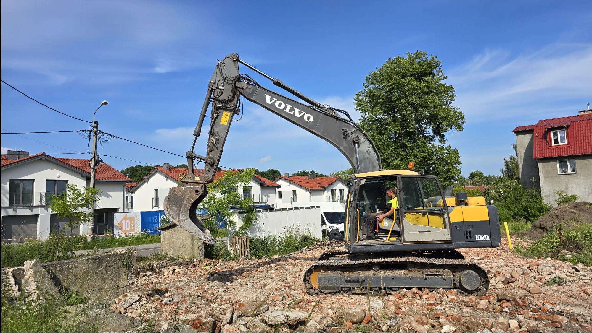 Żółto-szara koparka Volvo na gruzowisku, w tle zabudowa mieszkalna i błękitne niebo. Operator w kabinie. Widoczne fragmenty zburzonej konstrukcji i cegieł.