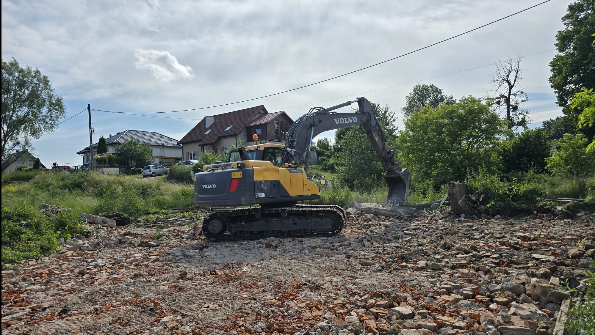 Żółto-szara koparka Volvo E160DL na terenie z gruzem i cegłami po rozbiórce, w tle budynki mieszkalne i zieleń.